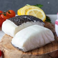 two pieces of fish on a cutting board with tomatoes and lemons in the background