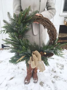 a woman is holding a wreath with pine cones and evergreens on it in the snow