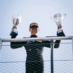 a man holding up two trophies while standing on top of a metal fence with his hands in the air