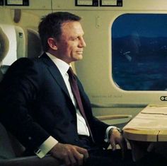 a man in a suit and tie sitting on an airplane looking out the window at something