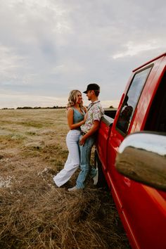 a man and woman leaning against the side of a red truck in an open field