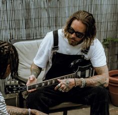 a man sitting on a bench playing an electric guitar with another person standing next to him