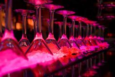 there are many wine glasses lined up on the bar counter with pink lights behind them