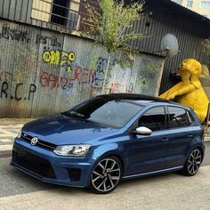 a blue volkswagen car parked in front of a graffiti covered building with a yellow bear statue behind it