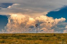 a windmill in the middle of an open field with clouds above it and grass below