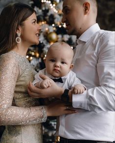 a man and woman holding a baby in front of a christmas tree with lights on it