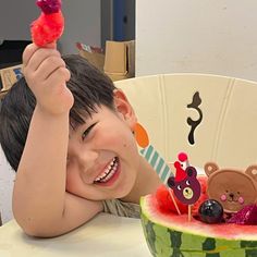 a young boy sitting at a table in front of a watermelon bowl holding up a piece of fruit