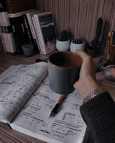 a person holding a coffee cup on top of an open book next to a pen