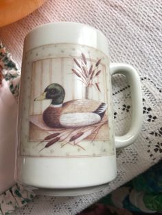 a coffee cup with a duck on it sitting on a tablecloth next to a vase