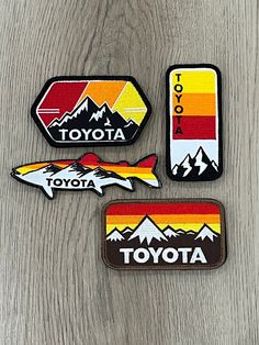 three patches with the words toyota and mountains in them on a wooden surface, one has a sticker that says totota