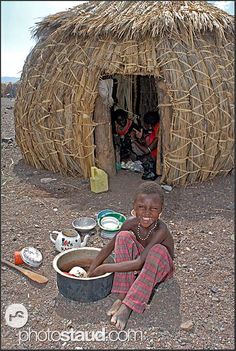 a young boy sitting in front of a hut with straw walls and flooring on the ground