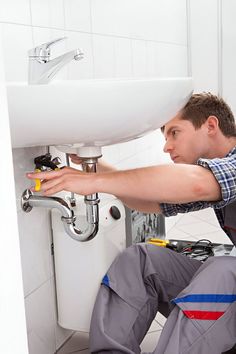 a plumber fixing a sink in a bathroom with white tiles on the wall behind him