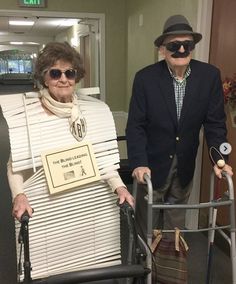 two elderly people with canes and hats in an office hallway, one pushing a walker