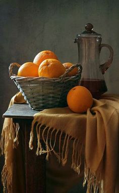 a basket of oranges sitting on top of a wooden table next to a pitcher