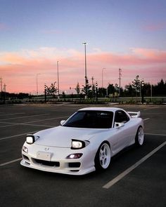 a white sports car is parked in a parking lot at dusk with the sun setting