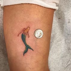 a woman's leg with a small mermaid tattoo on it, next to a penny