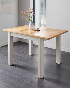 a white vase with flowers sitting on top of a wooden table next to a window