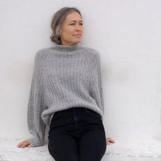 a woman sitting on the ground wearing black jeans and a gray sweater with her hands in her pockets