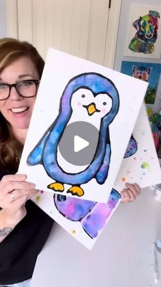 a woman holding up a painting with a penguin on it's face and smiling at the camera