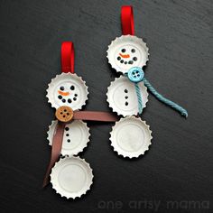 two snowmen made out of bottle caps on a black surface with red ribbon around them