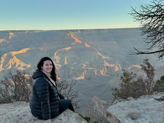 a woman sitting on the edge of a cliff at sunset with mountains in the background