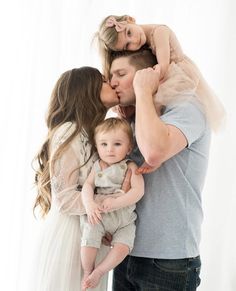 a man and woman holding a baby in front of a white background with the child's mother kissing her cheek