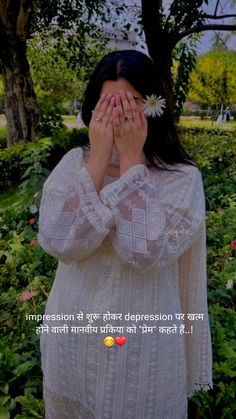 a woman covers her face with her hands while standing in front of trees and flowers