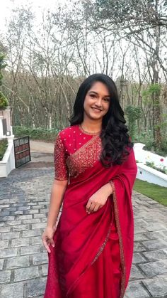 a woman in a red sari standing on a brick walkway with trees and bushes behind her