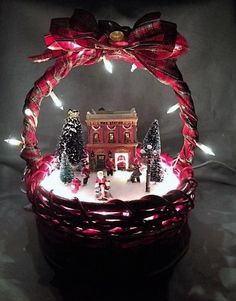 a lighted basket with a house and trees in it on top of a table next to some lights