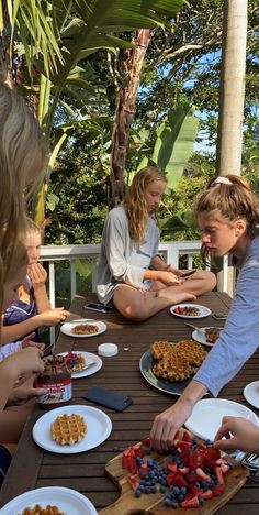 a group of people sitting around a table with plates of food on it and one person cutting up some fruit