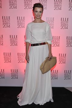 a woman standing in front of a pink wall with a purse on her hand and wearing a white dress