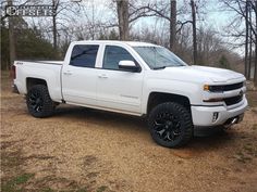 a white truck is parked in the dirt near some trees and bushes with black rims