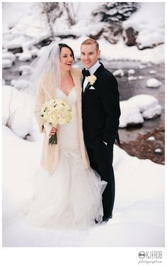 a bride and groom standing in the snow near a stream wearing tuxedo jackets