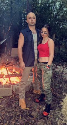 a man and woman standing in front of a campfire with their arms around each other