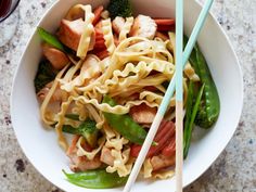 a white bowl filled with noodles, broccoli and meat next to chopsticks