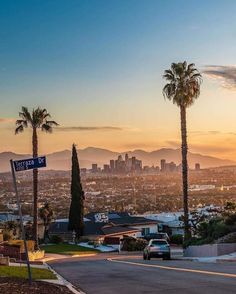 the sun is setting over palm trees and mountains in this cityscape with cars parked on the street