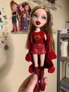 a hand holding a doll with long blonde hair and blue eyes in a red outfit