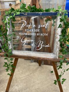 a wooden easer holding a glass sign with greenery on it and the words, pitt & seat either side you're loved by both the groom and bride