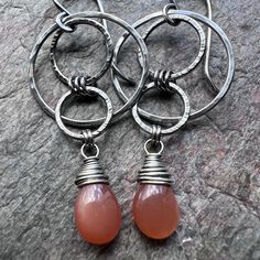 Natural Peach Moonstone teardrops have been wrapped in sterling silver wire.  The stones dangle below a set of sterling silver circles I have formed, soldered, and textured by hand.  The earrings dangle from handformed sterling silver earwires.  The silver has been oxidized and polished to create a patina finish. Be sure to see all photos and video for size and color reference.   Total Earring Length (Including Earwire): Just under 2 inches Total Earring Weight: About 2.1 grams per one earring A Modern Teardrop Wire Wrapped Jewelry, Modern Wire Wrapped Teardrop Jewelry, Silver Carnelian Drop Earrings, Silver Carnelian Jewelry With Ear Wire, Silver Carnelian Teardrop Jewelry, Silver Teardrop Carnelian Jewelry, Silver Teardrop Earrings With Natural Stones, Silver Dangle Teardrop Earrings With Natural Stones, Silver Wire Wrapped Long Drop Teardrop Earrings