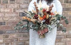 a woman holding a bouquet of dried flowers in front of a brick wall with foliage