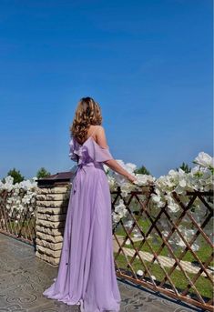 a woman in a long purple dress standing on a bridge looking at the sky and flowers