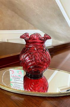 a red glass vase sitting on top of a wooden table next to a mirror plate