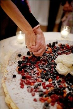 two people holding hands over a cake with berries and cream on it