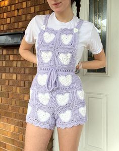 a woman standing in front of a brick wall wearing purple and white crocheted overalls