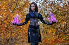 a woman dressed in black holding purple flowers with her hands out to the side, surrounded by trees and leaves