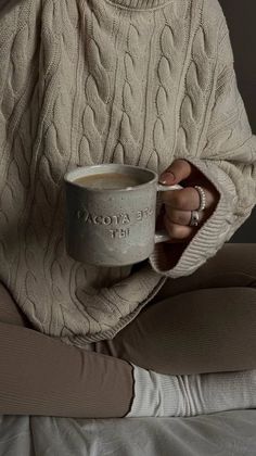 a woman holding a cup of coffee while sitting on a bed with her hands wrapped around the mug