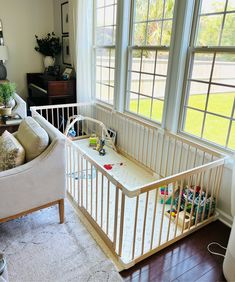 a baby crib in the corner of a living room with two windows and a couch