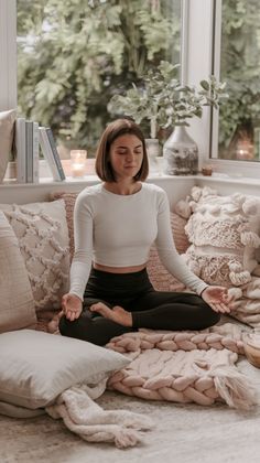 a woman sitting in a lotus position on a couch with pillows and blankets around her