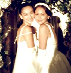 two young women in white dresses standing next to each other under a floral arch with greenery