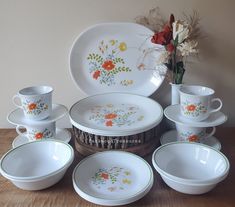 an assortment of white dishes and cups on a wooden table next to a vase with flowers